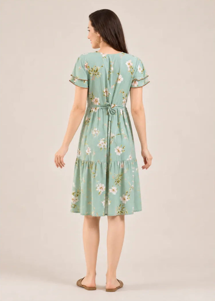 A woman viewed from the back wearing a light sage green midi dress with a white and pink floral print, short flutter sleeves, and a tie at the waist, set against a solid beige studio background.