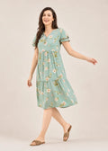 A woman modeling a light green tiered midi dress with a white and pink floral print, featuring short puff sleeves and a V-neckline, set against a plain beige studio background.