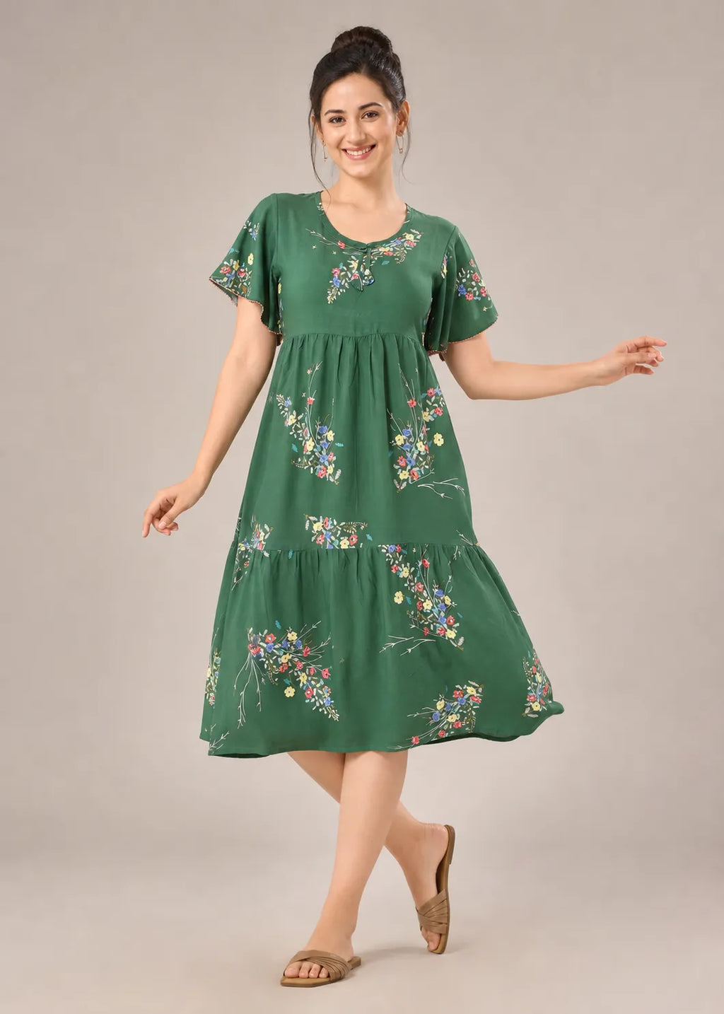 A woman modeling a green tiered midi dress with short sleeves and a colorful floral print, standing against a solid light gray studio background.