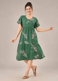 A woman modeling a green tiered midi dress with short sleeves and a colorful floral print, standing against a solid light gray studio background.