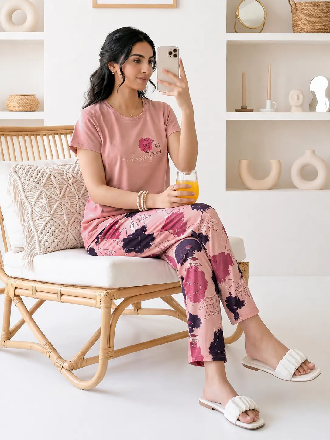 Woman in pink floral pajama set sitting on a chair, holding a phone and a glass.