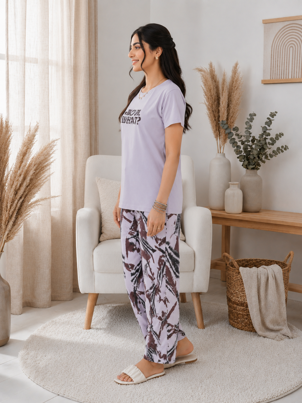 Woman in a light purple t-shirt and patterned pants standing in a bright living room.