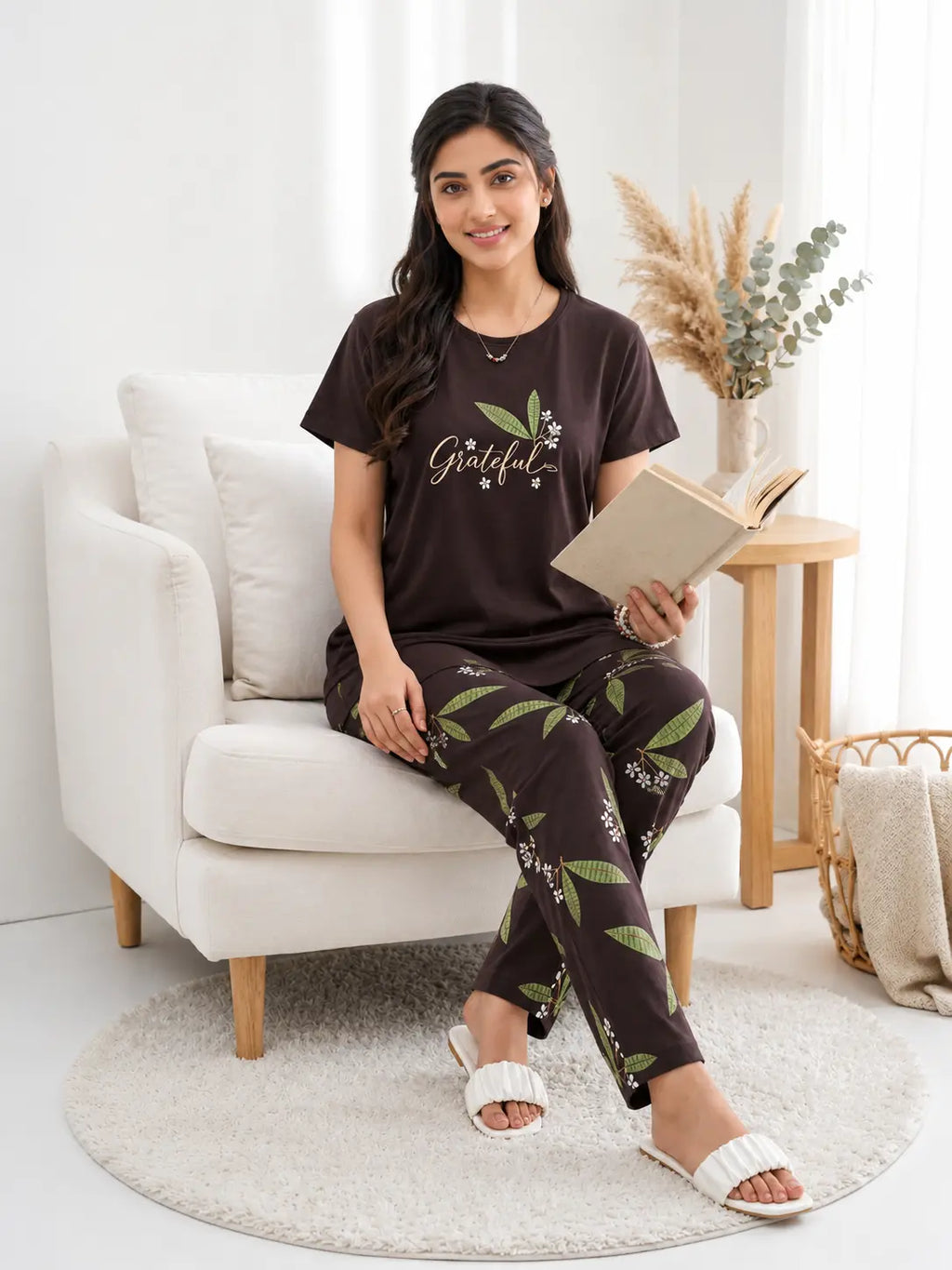 Woman sitting on a white armchair reading a book, wearing a 'Grateful' t-shirt and leaf-patterned pants.