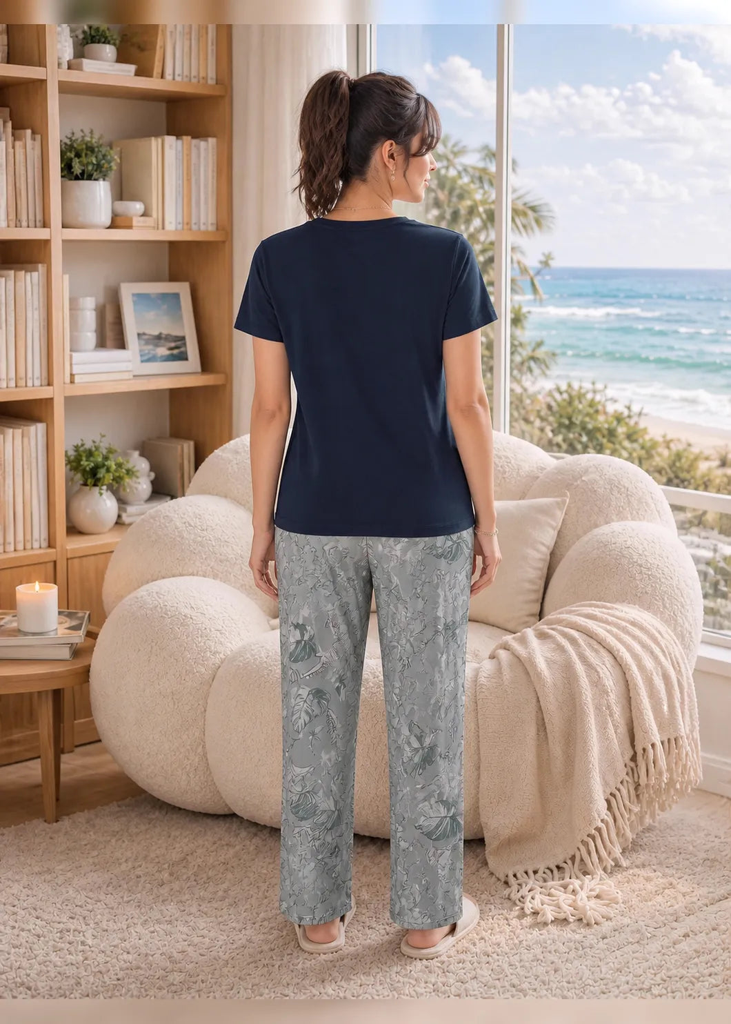 A woman stands with her back to the camera in a bright, modern room with a large window overlooking the ocean. She is wearing a navy blue short-sleeve top and light gray patterned pants.
