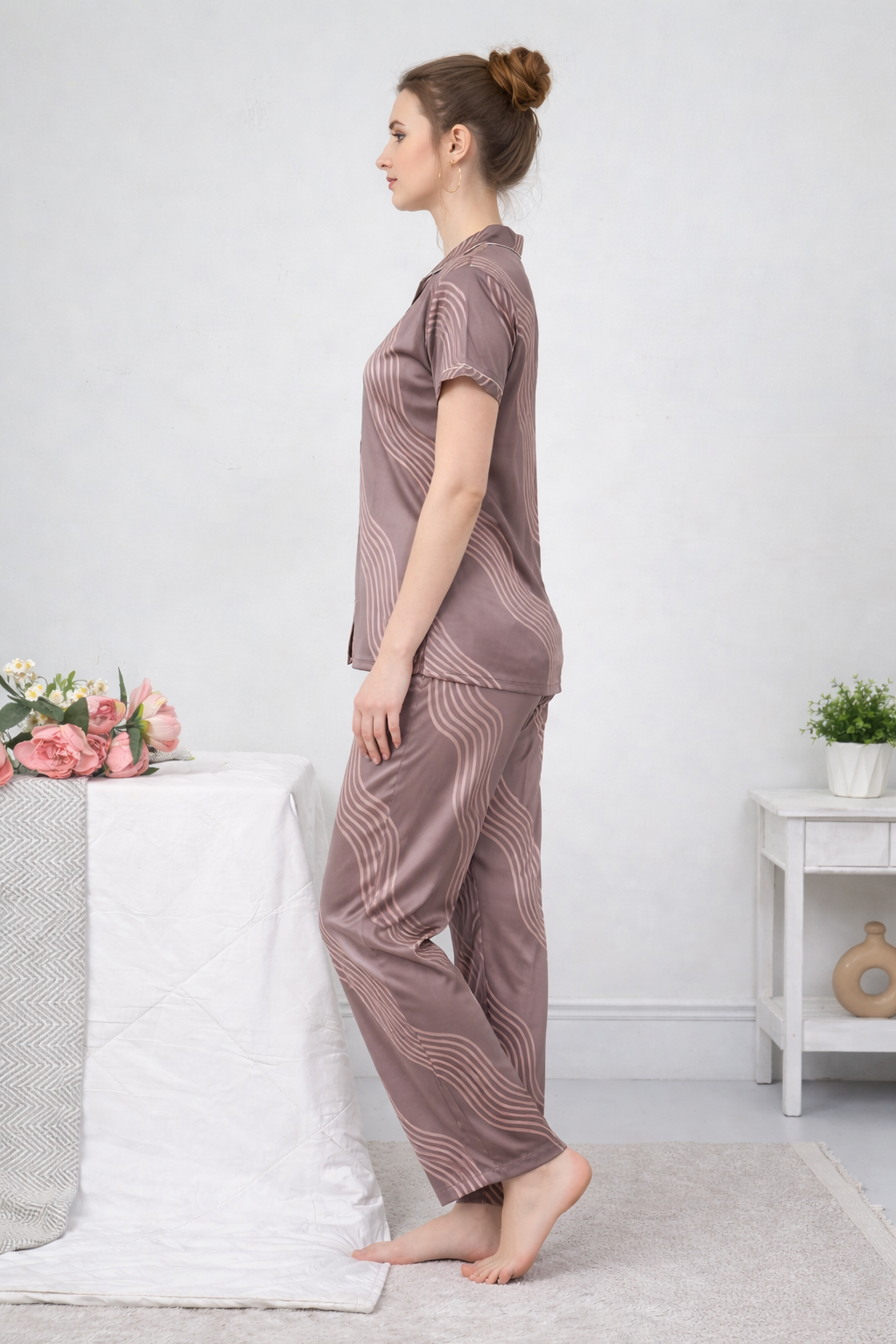 Woman wearing a matching pajama set in a room with a white bed and small table.