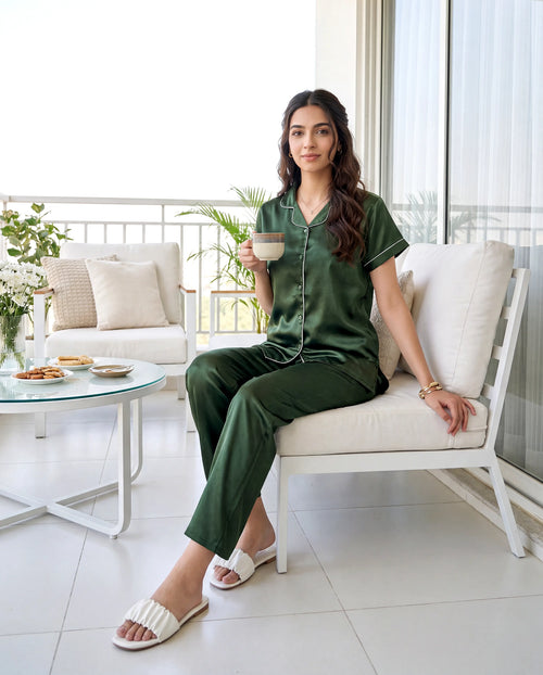 Woman in green pajama set sitting on a white chair holding a cup, with a modern living room in the background.