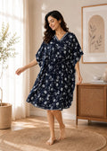 A woman modeling a navy blue kaftan dress with a white floral pattern in a warm, sunlit room with a wooden cabinet and framed art.