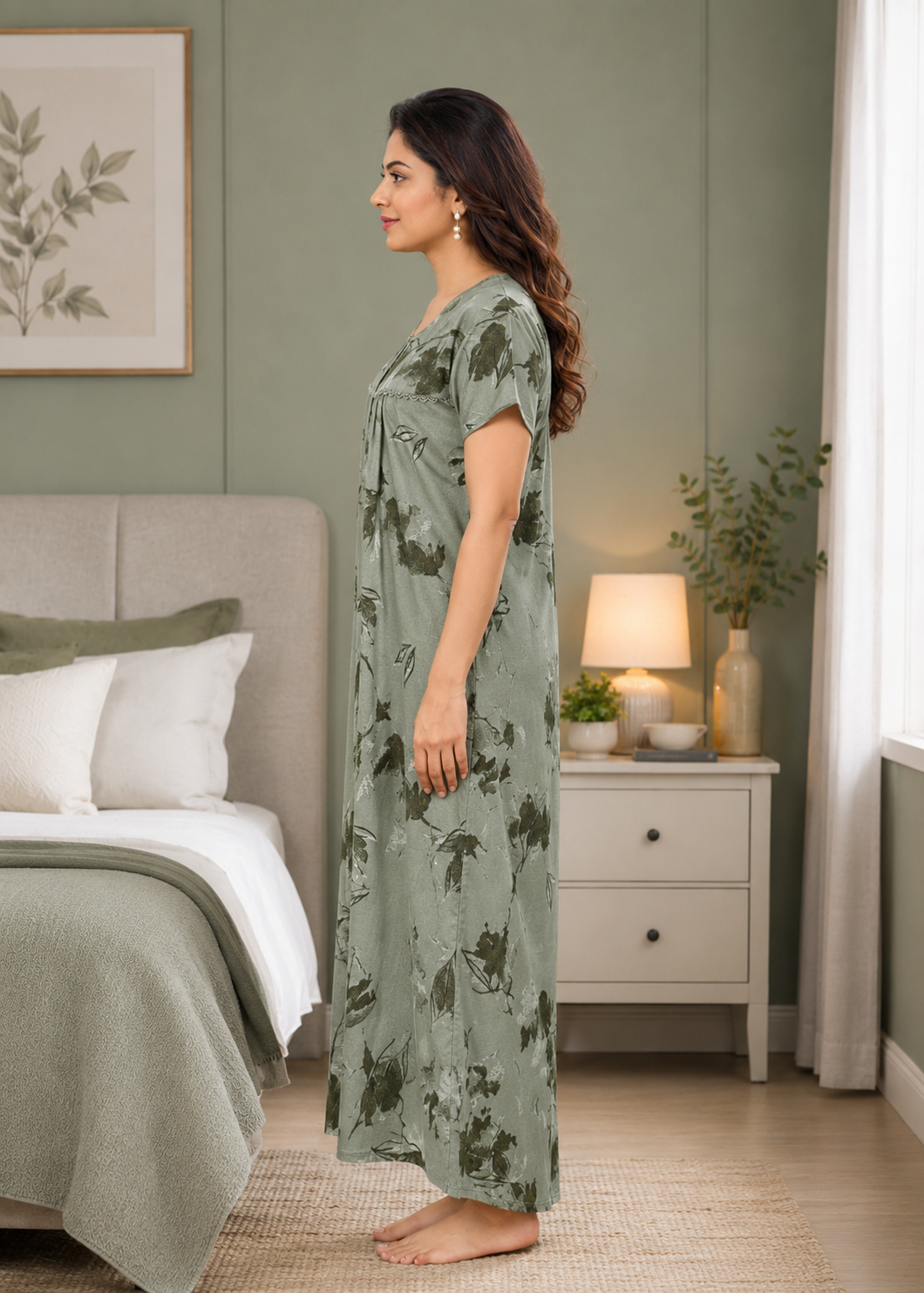 A woman stands in profile wearing a long sage green nightgown with a dark green floral print in a softly lit bedroom. The room features a bed, a white nightstand with a lit lamp, and a green wall.