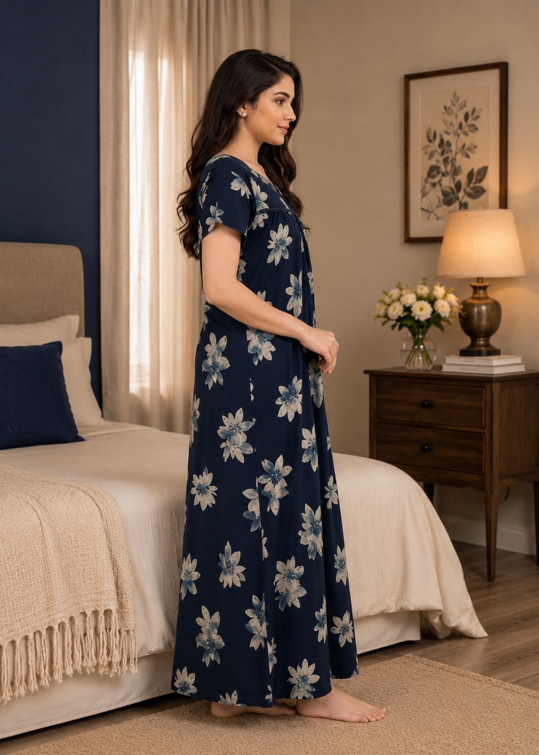 A woman stands in profile in a bedroom wearing a navy maxi dress with a white floral pattern. The room features a wooden nightstand with a lit lamp and flowers, a bed with cream bedding, and a dark blue accent wall.