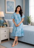 A woman wearing a light blue tiered nightgown with a darker blue floral pattern, standing in a bedroom setting with a bed, bedside table, and lamp in the background.