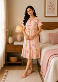 A woman wearing a light pink tiered nightgown with a floral print stands in a cozy bedroom setting next to a bed with a pink blanket.