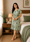 A woman stands barefoot in a bedroom wearing a light sage green tiered nightgown with a dark floral print. The room features a wooden nightstand with a lamp and plants, and a bed with white linens and a green throw blanket.