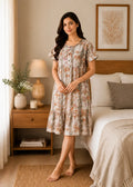 A woman stands barefoot in a bedroom wearing a short-sleeve midi-length nightgown with a soft floral print in muted beige, gray, and peach tones. The room includes a bed, wooden nightstand, lamp, and framed wall art.