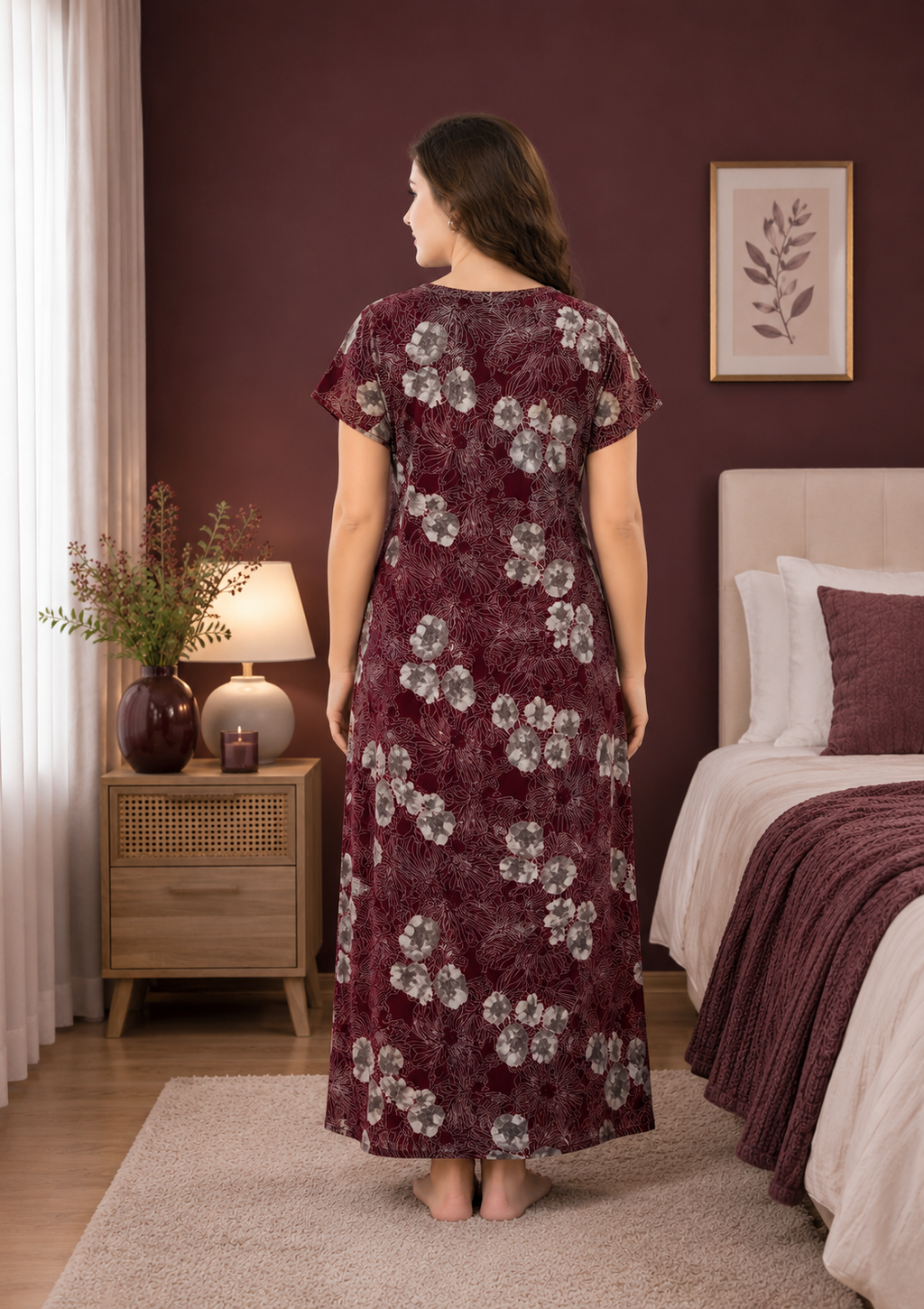 A woman stands with her back to the camera wearing a long-sleeved, floor-length nightgown with a floral pattern in deep burgundy and gray. She is photographed in a bedroom setting with a matching burgundy wall, a bed, and a bedside table.