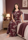 A woman wearing a long-sleeved maroon floral nightgown sits on the edge of a bed in a bedroom setting.