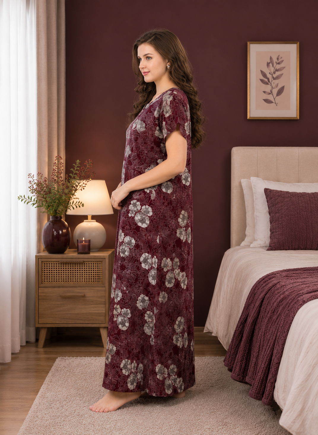A woman stands barefoot in a bedroom wearing a long-sleeved, floor-length maroon nightgown with a white floral pattern. The bedroom features a dark burgundy wall, a wooden bedside table with a lamp and vase, and a bed with matching bedding.