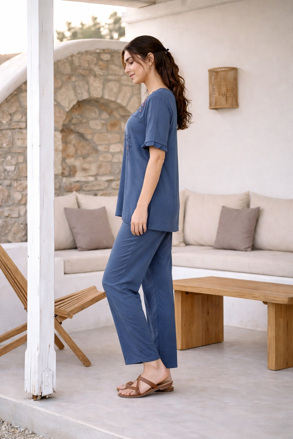 Woman in a blue outfit standing in a modern outdoor living space with stone walls and wooden furniture.