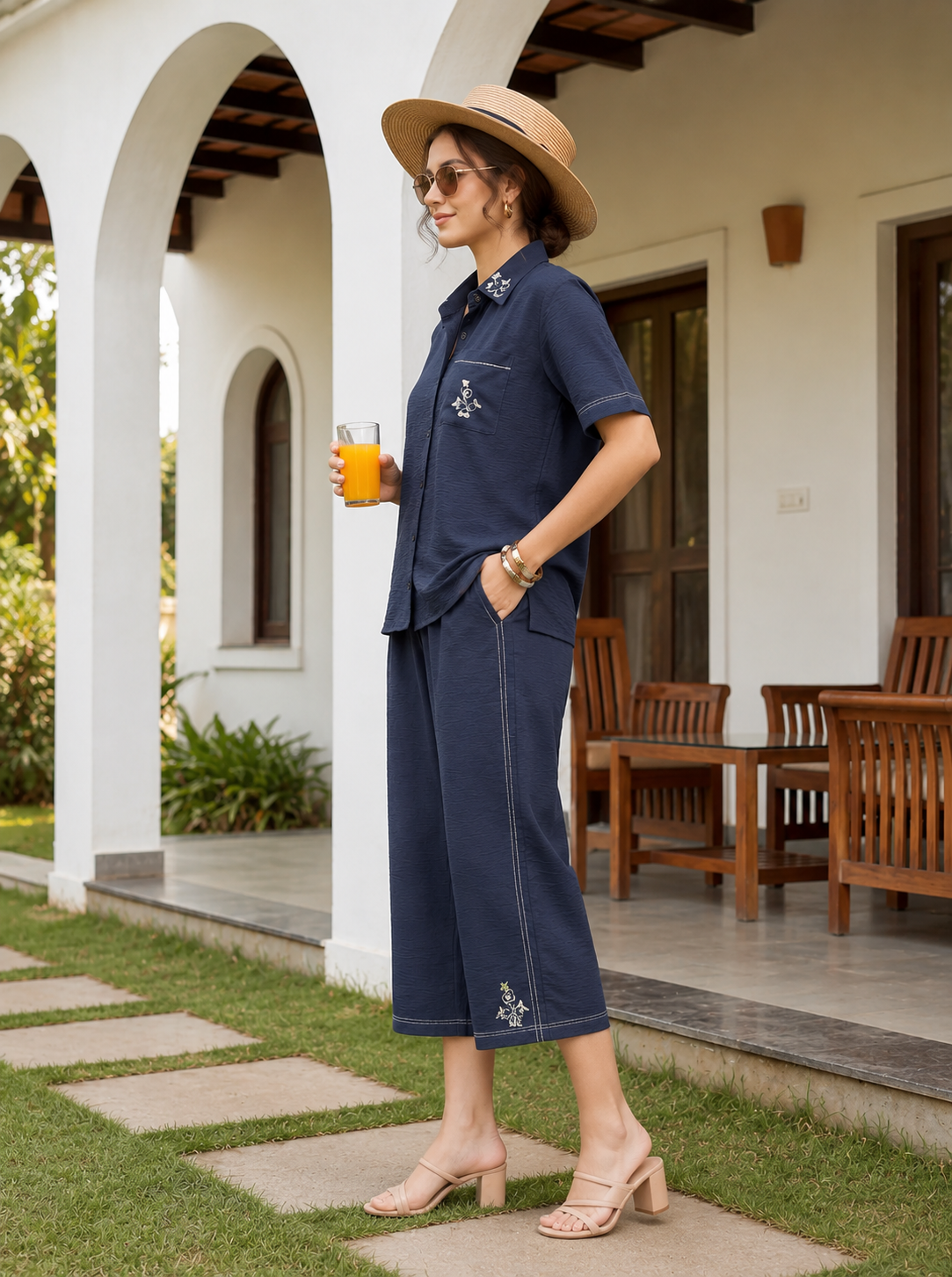 Woman in navy outfit with hat holding a drink, standing in front of a white building with wooden furniture.