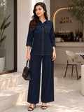 A woman models a dark navy blue two-piece jumpsuit set consisting of a long-sleeve button-up shirt and matching wide-leg trousers, styled with a black handbag and black sandals in a cafe setting.