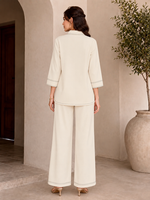 A woman stands with her back to the camera wearing a matching cream-colored two-piece lounge set consisting of a cropped jacket with three-quarter sleeves and wide-leg trousers. She is holding a small beige handbag and wearing beige high-heeled sandals in a warm, neutral-toned indoor setting with a potted plant.