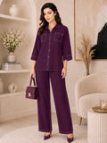 A woman models a matching deep purple two-piece lounge set consisting of a button-up shirt with three-quarter sleeves and wide-leg trousers, styled with a matching handbag and heels in a modern, neutral-toned living room setting.